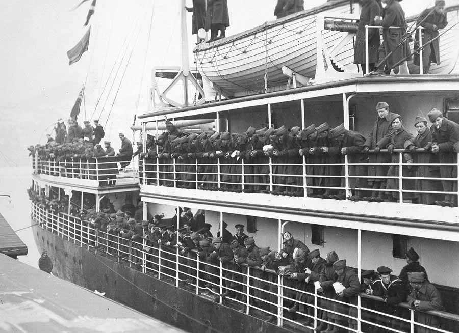 Black and white photograph. Hundreds of men in uniform solemnly lean over the rails of a ship as it sets sail from Newfoundland to England.